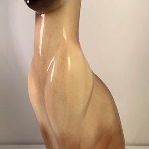 Op de afbeelding: Een keramieken Siamese kattenbeeldje in zittende houding. De kat heeft een glanzende afwerking met een kleurengradiënt van lichtbruin naar donkerbruin. De ogen zijn zwart en de neus en oren zijn donkerbruin.