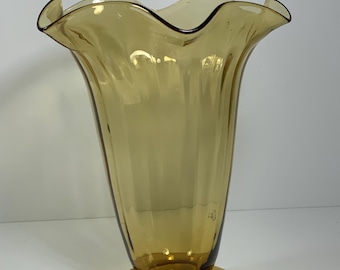 May include: Amber-colored glass vase with a flared, ruffled rim and a pedestal base. The vase has a tall, slender body with vertical ridges. The base is a circular shape. The glass has a warm, golden hue.