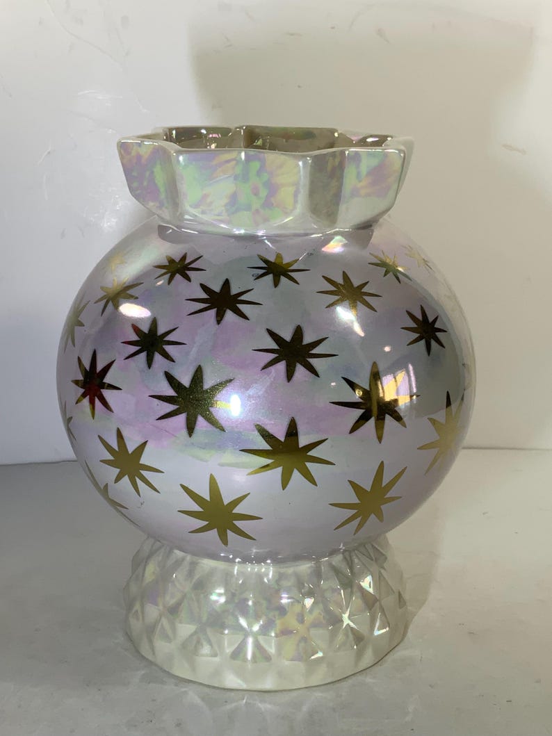 May include: Iridescent ceramic vase with a spherical body and a faceted base. The vase is decorated with gold starburst patterns. The top has a scalloped edge. The vase has a pearlescent finish that reflects multiple colors.