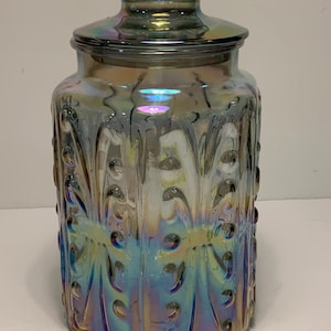 Op de afbeelding: Een iriserende glazen pot met een deksel. De pot heeft een decoratief patroon en een zeshoekige basis. De kleuren verschuiven van blauw naar goud en paars. Het deksel heeft een kleine, platte bovenkant met een reeks kleine gaatjes.