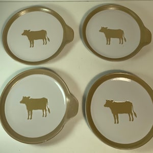 Puede incluir: Cuatro platos redondos blancos con borde dorado y silueta de vaca dorada en el centro. Cada plato tiene un asa dorada en el lateral. Los platos están dispuestos en forma cuadrada.