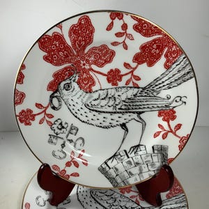 May include: Two white ceramic plates with a gold rim. The plates feature a black and white bird design with red floral accents. The plates are stacked on a wooden stand.