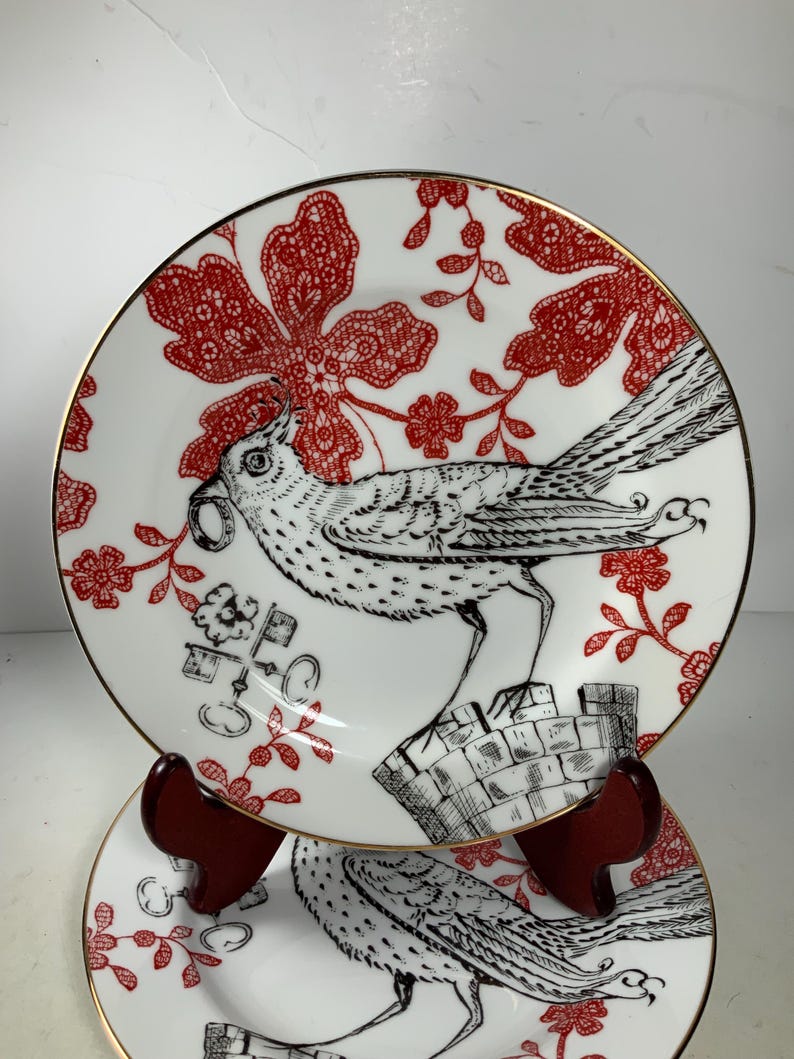 May include: Two white ceramic plates with a gold rim. The plates feature a black and white bird design with red floral accents. The plates are stacked on a wooden stand.