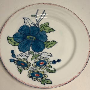 May include: White ceramic plate with a blue floral design. The plate features a large blue flower with green leaves and smaller blue and yellow flowers. The rim is decorated with a red scalloped pattern.