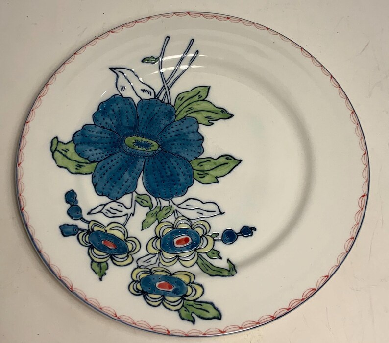 May include: White ceramic plate with a blue floral design. The plate features a large blue flower with green leaves and smaller blue and yellow flowers. The rim is decorated with a red scalloped pattern.