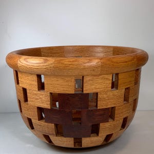 May include: A handcrafted wooden bowl with a woven design. The bowl is made of light and dark brown wood, with a geometric pattern of cut-out rectangles. The rim is rounded, and the bowl has a wide, open shape.