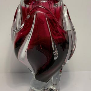 May include: A decorative glass vase with a swirling design. The vase features a combination of clear and deep red glass, creating a striking visual effect. The vase has a unique, twisted shape.