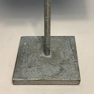 May include: A weathered, square metal base with a vertical rod extending upwards. The base is a dull gray color with a textured surface. The rod is also gray and appears to be made of the same material. The object is likely a stand or support.