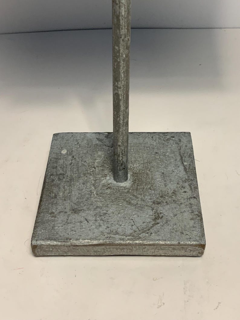 May include: A weathered, square metal base with a vertical rod extending upwards. The base is a dull gray color with a textured surface. The rod is also gray and appears to be made of the same material. The object is likely a stand or support.