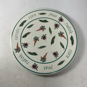 May include: Round white ceramic trivet with a green border and a pattern of peas, carrots, corn, and radishes. The words "corn", "peas", "radish", "corn", "radish", and "peas" are written around the border.