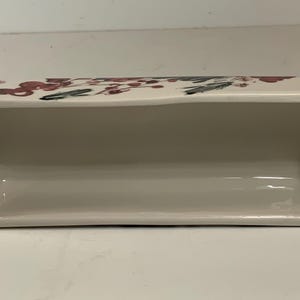 May include: Rectangular ceramic dish with a cream-colored interior and exterior. The top edge features a floral design with red and green accents. The dish is ideal for serving or display.