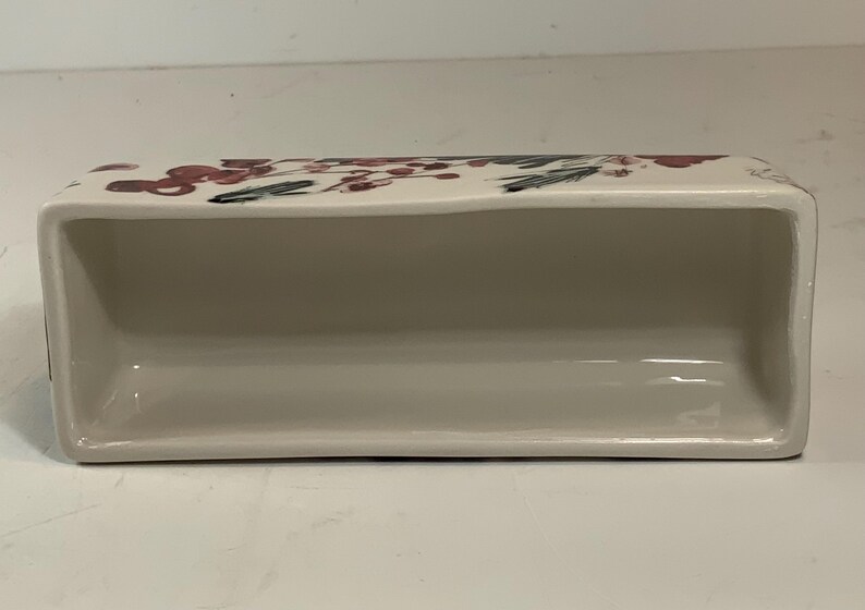 May include: Rectangular ceramic dish with a cream-colored interior and exterior. The top edge features a floral design with red and green accents. The dish is ideal for serving or display.