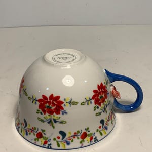 May include: White ceramic teacup with a blue handle and floral design. The cup features a vibrant pattern of red flowers, green leaves, and accents of yellow and blue. A small, pink floral accent is on the handle.