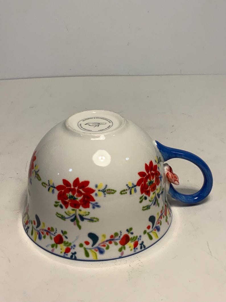 May include: White ceramic teacup with a blue handle and floral design. The cup features a vibrant pattern of red flowers, green leaves, and accents of yellow and blue. A small, pink floral accent is on the handle.