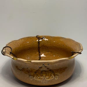 May include: A ceramic hanging planter with a scalloped rim and a brown glaze. The planter has a decorative embossed design and is suspended by a metal chain. The planter is approximately 8 inches in diameter.