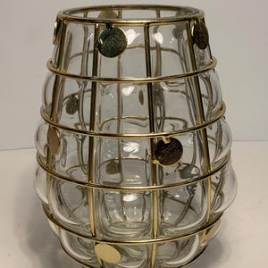 Op de afbeelding: Een heldere glazen vaas met een afgeronde vorm, omhuld door een goudkleurige metalen kooi. De kooi heeft verticale en horizontale staven, met gouden schijfaccenten. De vaas heeft een decoratief, modern design.