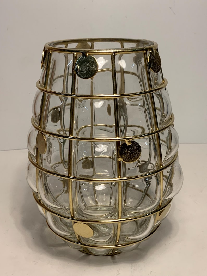 Op de afbeelding: Een heldere glazen vaas met een afgeronde vorm, omhuld door een goudkleurige metalen kooi. De kooi heeft verticale en horizontale staven, met gouden schijfaccenten. De vaas heeft een decoratief, modern design.