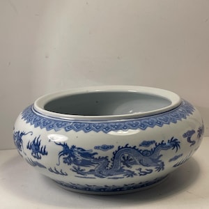 Puede incluir: Un cuenco redondo de cerámica blanca con un diseño de dragón y nubes azules. El cuenco tiene una abertura ancha y un borde decorativo. El dragón está representado de forma detallada y estilizada, con escamas y líneas fluidas. El cuenco es probablemente para fines decorativos.