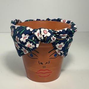 May include: A terracotta flower pot with a painted face, adorned with a blue floral headband. The headband features white flowers with pink centers and green leaves. The face is painted with blue eyes and red lips.
