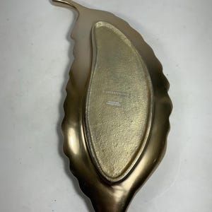 Puede incluir: Una bandeja de servir dorada en forma de hoja con un borde elevado y un interior liso y plano. La bandeja est&aacute; hecha de metal y tiene una superficie ligeramente texturizada. La parte posterior de la bandeja est&aacute; estampada con el texto "Anthropologie" y "Hecho en India".