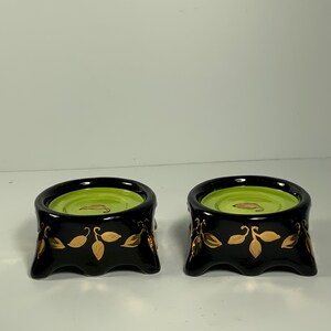 Può includere: Due portacandele in ceramica nera con dettagli in verde e oro. I portacandele hanno una parte superiore rotonda e una base con quattro piedi.