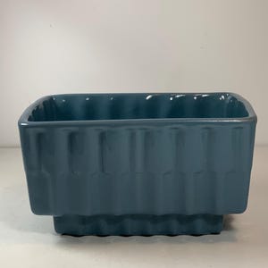 May include: A rectangular, teal-colored ceramic planter with a ribbed design on the sides. The planter has a slightly raised base and a glossy finish. The interior is smooth and the corners are rounded.
