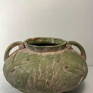 May include: A large, green ceramic vase with two handles and a wide opening. The vase features embossed floral and leaf designs. The exterior has a weathered appearance, with hints of pink and brown peeking through the green glaze.