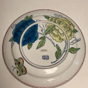 May include: White ceramic plate with a blue and yellow floral design, green leaves, and a red border. A butterfly is on the side. The plate is marked "Made in China" and "Molly Hatch."