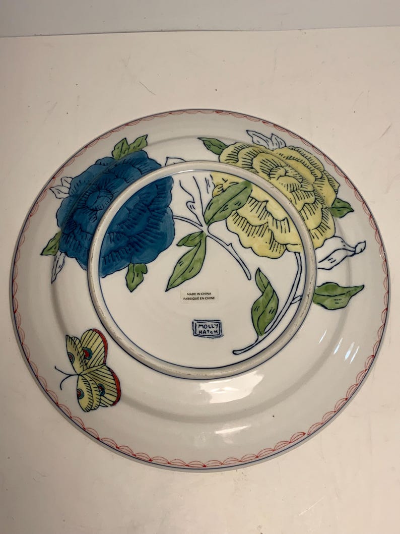 May include: White ceramic plate with a blue and yellow floral design, green leaves, and a red border. A butterfly is on the side. The plate is marked "Made in China" and "Molly Hatch."