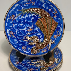 May include: Set of four decorative plates with a blue background and a gold rim. Each plate features a detailed dragon design in shades of brown, orange, and green. The plates have a glossy finish and are stacked on a black stand.