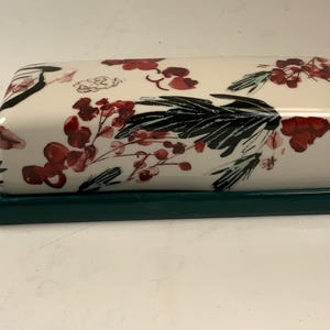 May include: A rectangular butter dish with a cream-colored lid and a dark green base. The lid is decorated with a floral pattern featuring red berries and black leaves. The dish is designed for kitchen use.