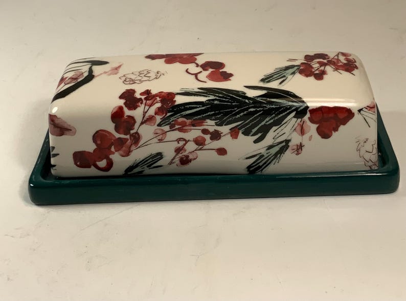 May include: A rectangular butter dish with a cream-colored lid and a dark green base. The lid is decorated with a floral pattern featuring red berries and black leaves. The dish is designed for kitchen use.