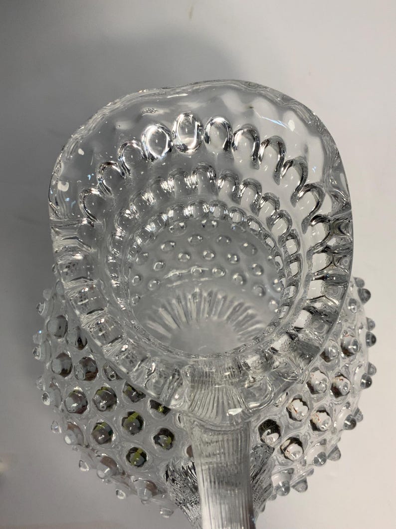May include: Clear glass pitcher with a hobnail design. The pitcher has a scalloped rim and a handle. The hobnail pattern covers the body of the pitcher. The pitcher is empty and appears to be vintage.