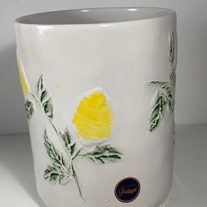 May include: White ceramic vase with embossed yellow lemons and green leaves. The vase has a slightly curved shape and a label that says "Handcrafted in Portugal". The design features a bright yellow color.