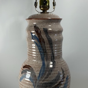 May include: A ceramic table lamp with a beige speckled finish and blue and brown abstract brushstrokes. The lamp has a brass-coloured metal frame and a clear lightbulb. The lamp is a decorative home decor item.