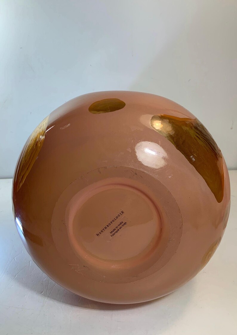 May include: A peach-colored, round decorative vase with gold accents. The vase has a glossy finish and a wide opening. The bottom of the vase has a label that reads "by ANTHROPOLOGIE Made in India Fabriqu&eacute; en Inde."