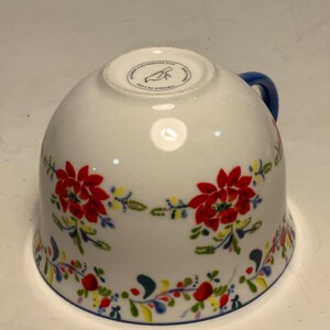 May include: White ceramic teacup with a blue handle and a floral design. The cup features red flowers, green leaves, and accents of yellow and blue. A blue line trims the rim.