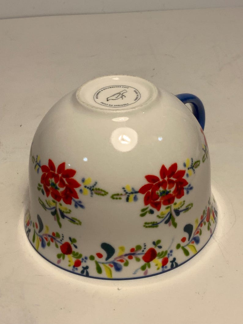 May include: White ceramic teacup with a blue handle and a floral design. The cup features red flowers, green leaves, and accents of yellow and blue. A blue line trims the rim.