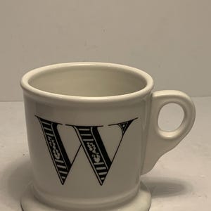 Puede incluir: Taza de cer&aacute;mica blanca con una gran letra "W" negra en la parte delantera. La letra est&aacute; decorada con patrones de rayas y flores. La taza tiene un asa peque&ntilde;a y una base ancha.