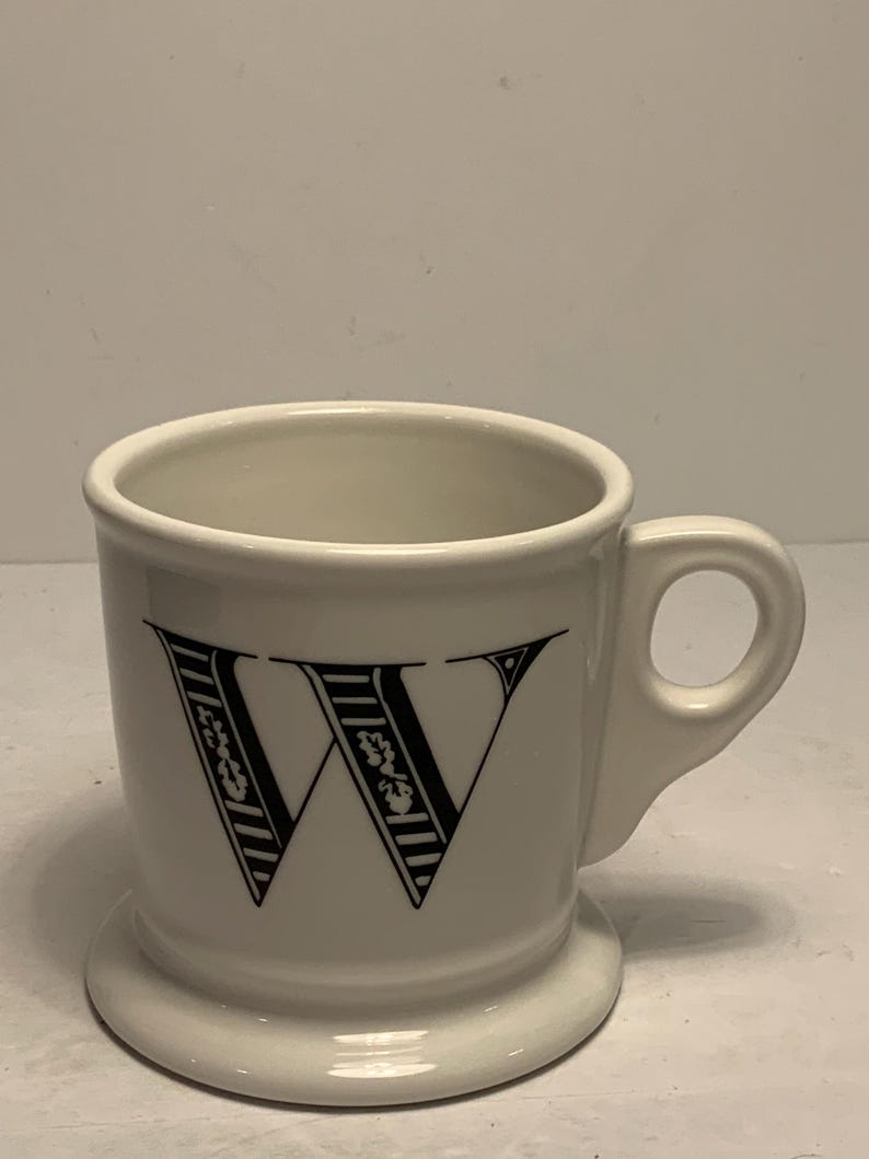 Puede incluir: Taza de cer&aacute;mica blanca con una gran letra "W" negra en la parte delantera. La letra est&aacute; decorada con patrones de rayas y flores. La taza tiene un asa peque&ntilde;a y una base ancha.