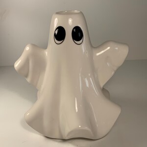 May include: A white ceramic ghost-shaped candle holder with large black eyes and a smiling expression. The ghost has outstretched arms and a draped body, perfect for Halloween decor. The candle holder is approximately 6 inches tall.