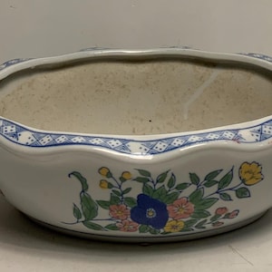 May include: White ceramic planter with a scalloped rim and floral design. The planter features a blue and white patterned border and a colorful floral motif with blue, pink, and yellow flowers.