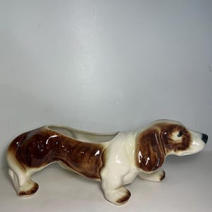 May include: A ceramic planter shaped like a long-bodied dog with a glossy finish. The dog features a white base coat with brown patches on the ears, back, and hindquarters, and a black nose. The open top of the planter is located on the back.