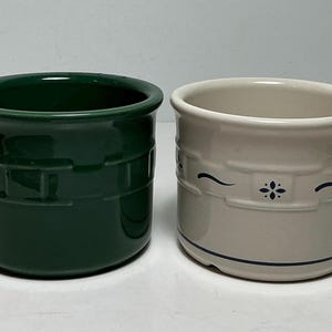 May include: Two cylindrical ceramic crocks with a raised basket-weave texture around the exterior. One crock is solid dark forest green, while the other is off-white with a dark blue floral motif and thin horizontal line accents near the base.