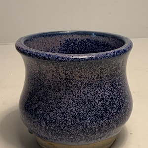 May include: A handmade ceramic vase with a speckled blue glaze. The vase has a rounded body and a flared rim. The base is a natural tan color. The interior is a darker blue. This decorative piece is suitable for holding flowers or as a standalone art object.