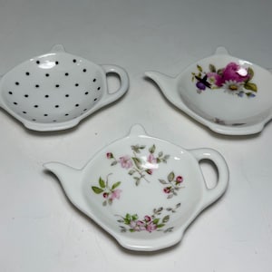 Könnte beinhalten: Drei weiße Teebeutelablagen aus Keramik in Form von Miniatur-Teekannen. Eine hat ein schwarzes Punktemuster, eine andere zeigt ein florales Design mit rosa Rosen und Gänseblümchen, und die dritte ist mit kleinen rosa Blüten und grünen Blättern verziert.