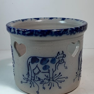 Puede incluir: Un jarrón de cerámica gris claro con un borde azul oscuro e ilustraciones de vacas. El jarrón tiene recortes en forma de corazón y un diseño detallado de vaca. La vaca tiene manchas azul oscuro y está sobre hierba azul.