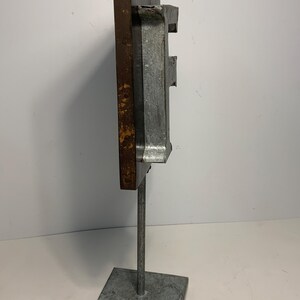 May include: A metal sculpture with a rectangular, weathered brown and gray form mounted on a square base. The sculpture has a vertical orientation and appears to be made of industrial materials. The base is square and the support is a thin metal rod.