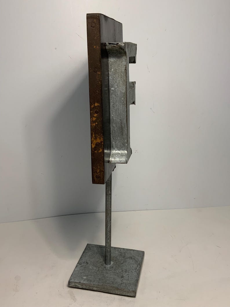 May include: A metal sculpture with a rectangular, weathered brown and gray form mounted on a square base. The sculpture has a vertical orientation and appears to be made of industrial materials. The base is square and the support is a thin metal rod.