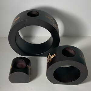 Puede incluir: Conjunto de tres candelabros de madera negra con dise&ntilde;os florales dorados. Los candelabros son de diferentes tama&ntilde;os y formas, con una abertura redonda en el centro de cada uno.